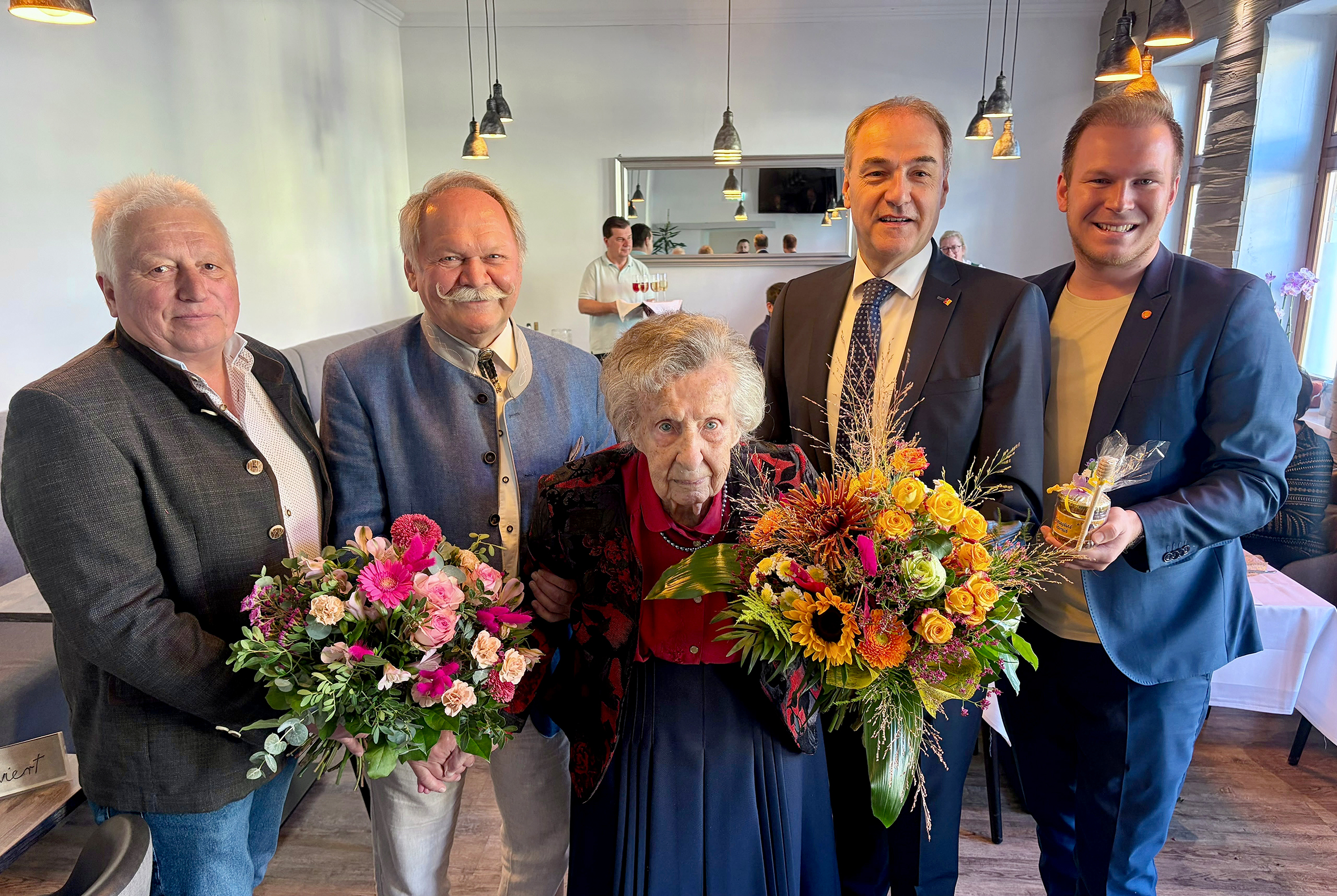 Jubilarin Hedwig Weinhofer feiert ihren 100. Geburtstag in Heiligenkreuz im Lafnitztal, umringt von Gratulanten mit Blumensträußen, darunter Soziallandesrat Leonhard Schneemann